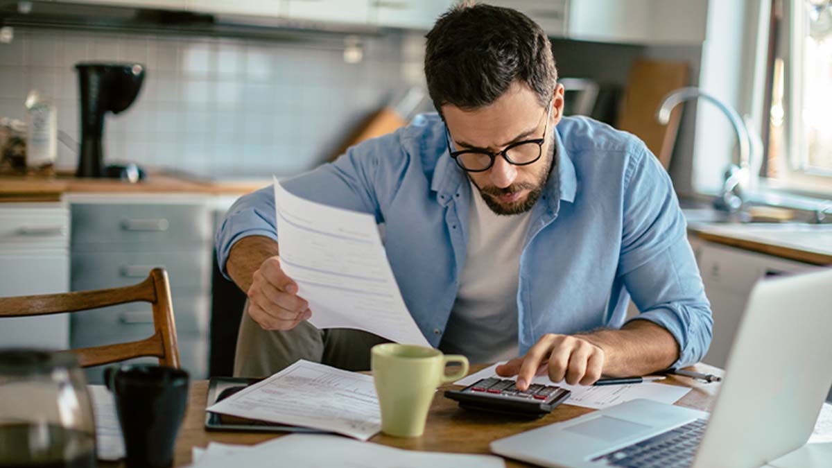 Homem a verificar as duas contas com calculadora, portátil e documentos numa mesa